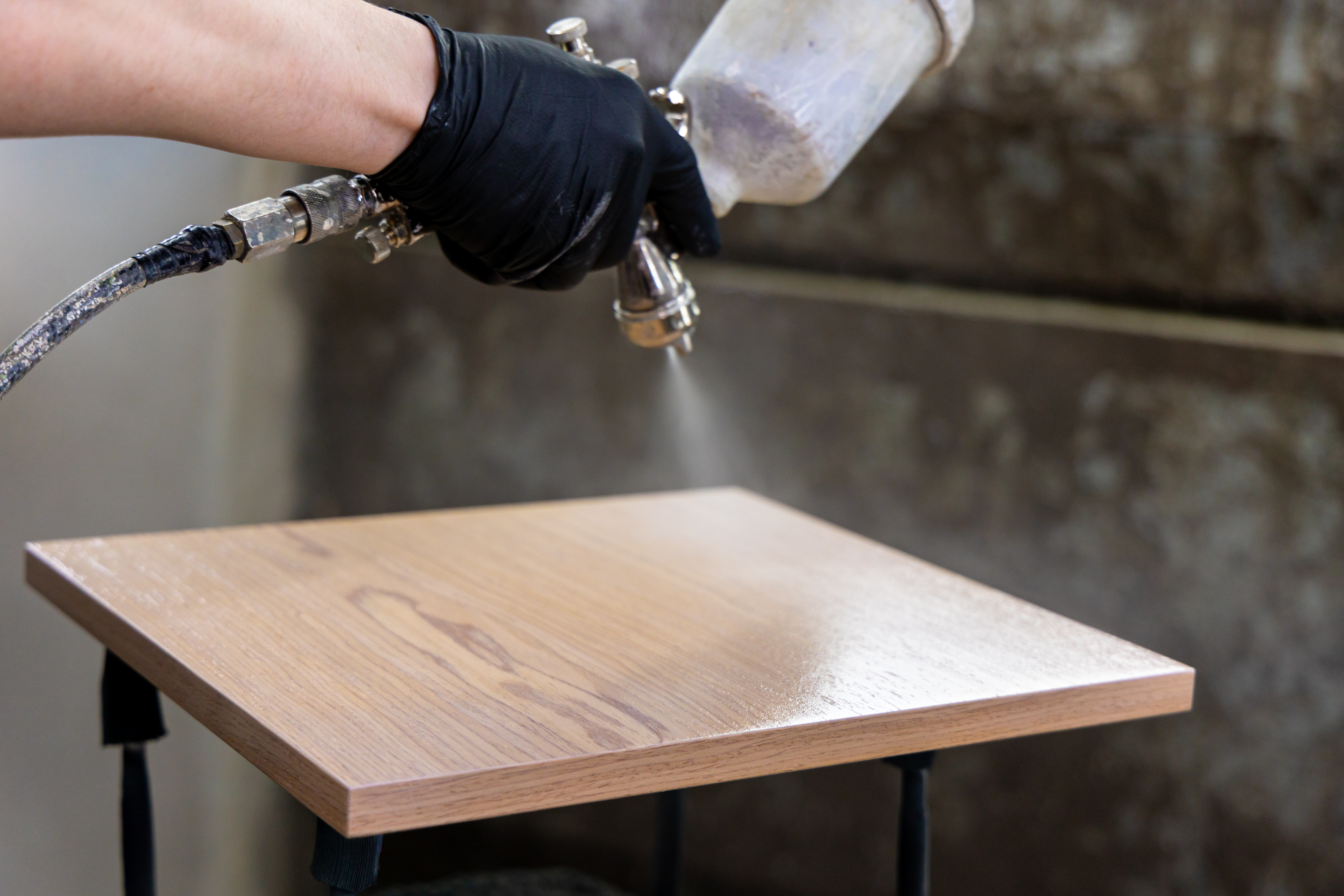 Worker spraying protective coating on wooden panel with spray gun. Furniture finishing process in workshop.